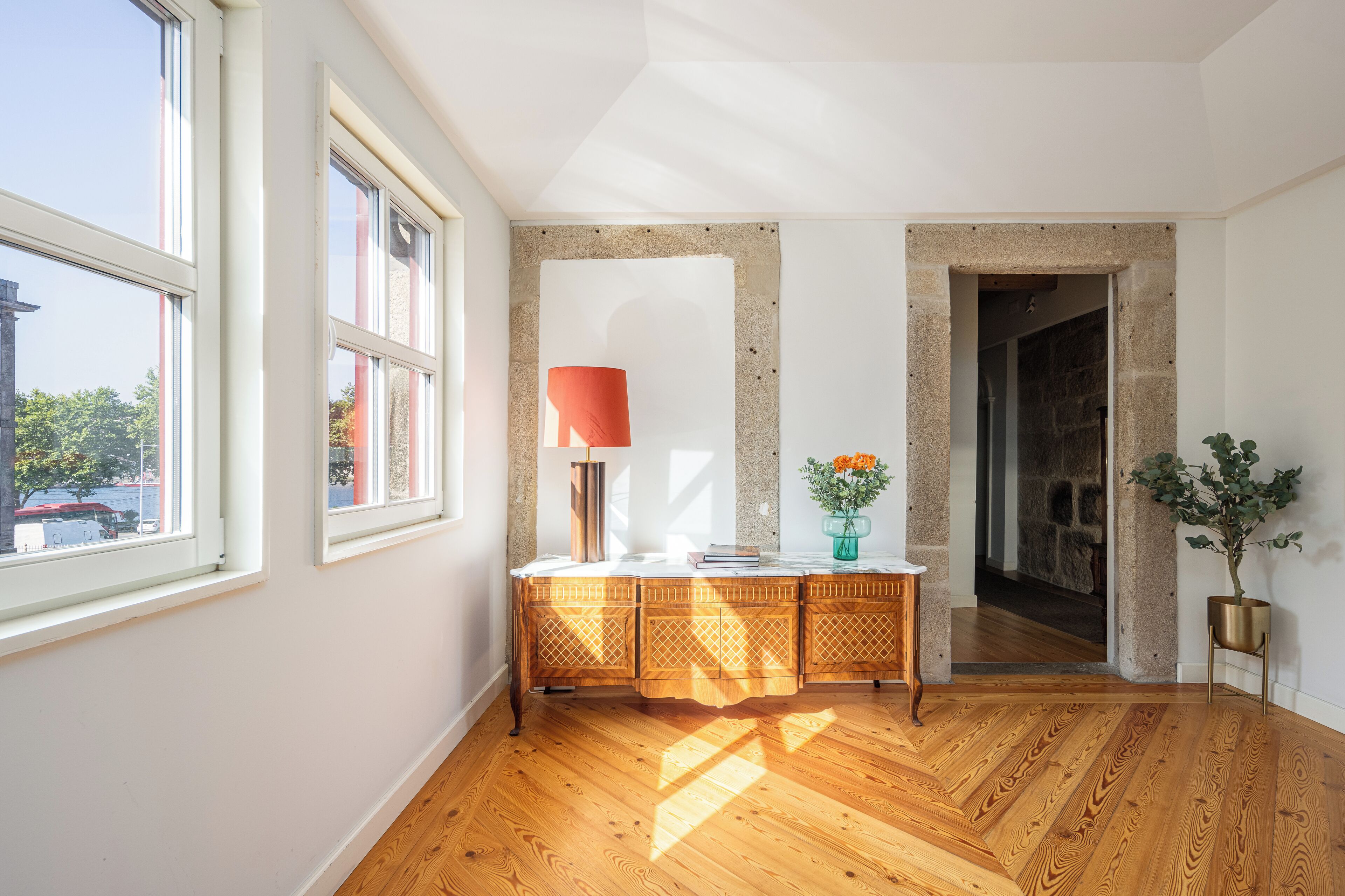 A room with wooden flooring, a white wall, a window, a red lamp, a vase with flowers, and a potted plant.
