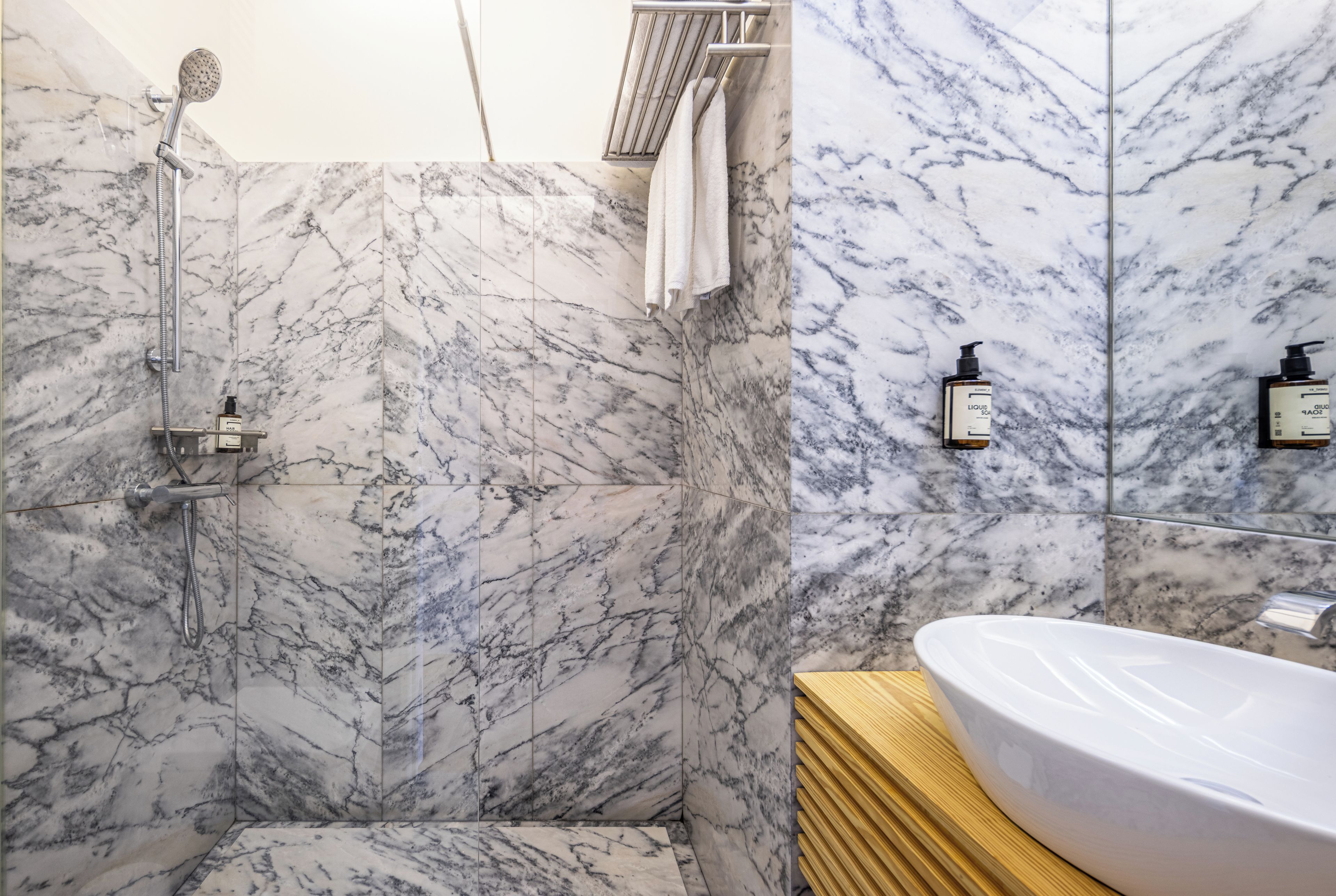 Un baño con paredes de mármol, ducha, lavamanos y artículos de aseo.