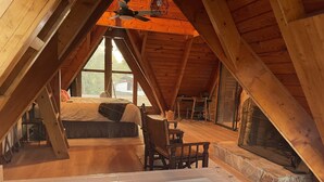 Een zolderkamer met een groot raam, houten plafond, een bed, een stoel en een open haard.