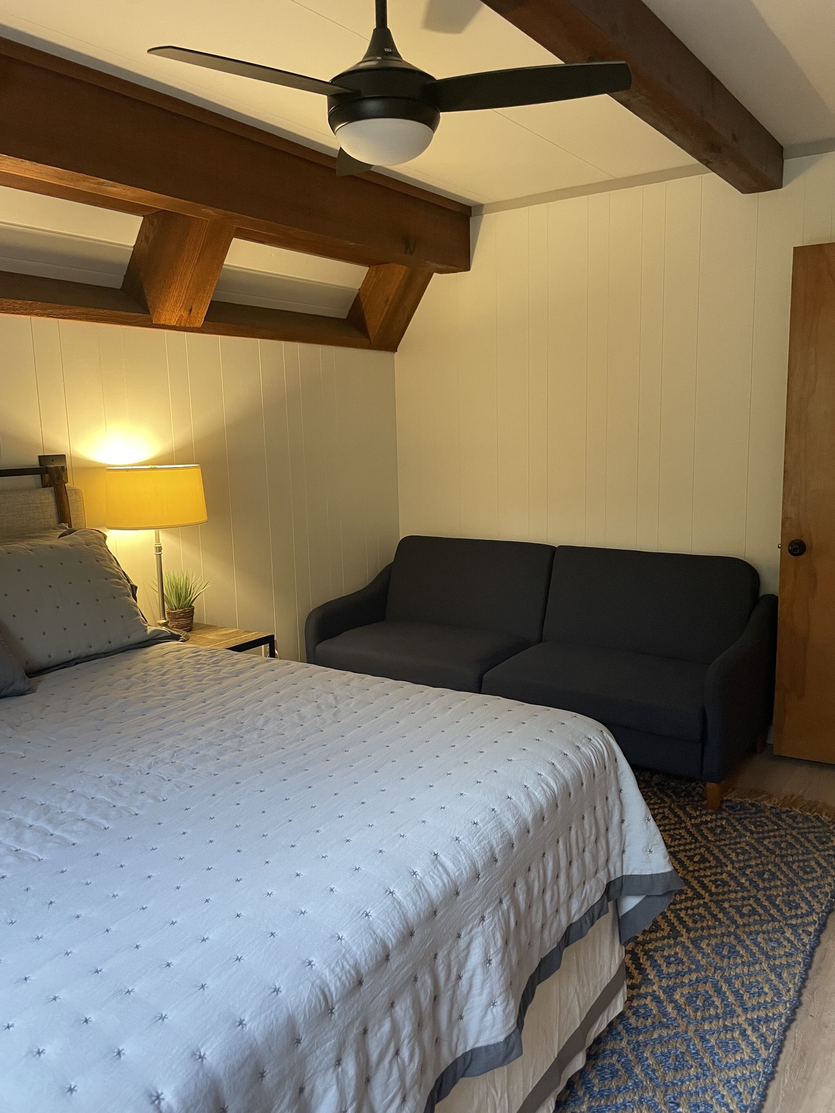 A bedroom with a bed, a dark sofa, a ceiling fan, a lamp, and a wooden door.