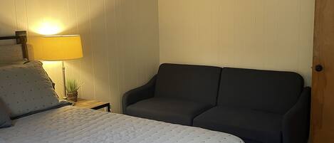 Un dormitorio con una cama, un sofá oscuro, un ventilador de techo, una lámpara y una puerta de madera.