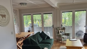 A spacious living room with a green sofa, a white sofa, and a wooden coffee table. There is a dining table with chairs, a plaid armchair, and a large window with a view of trees and a car. - Stunning 1 Bedroom Lodge (Welshpool)