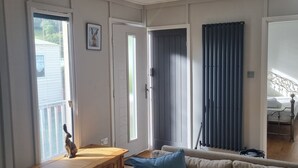 A living room with a wooden bench, a beige sofa with a blue pillow, a door, and a window with blinds. - Stunning 1 Bedroom Lodge (Welshpool)