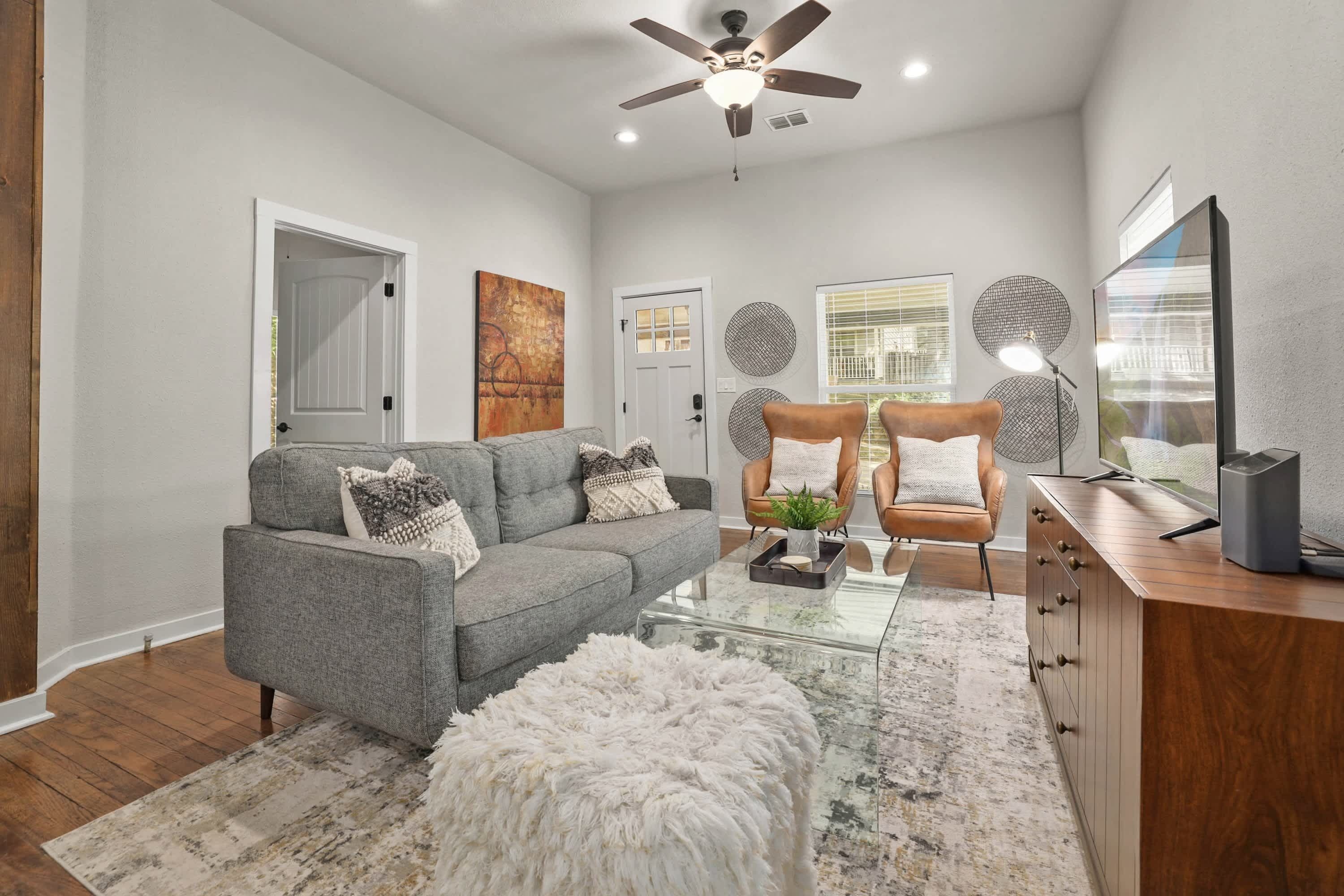 A living room with a grey sofa, a white fluffy ottoman, a glass coffee table, and a wooden cabinet with a TV.