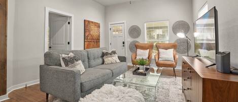 A living room with a grey sofa, a white fluffy ottoman, a glass coffee table, and a wooden cabinet with a TV.