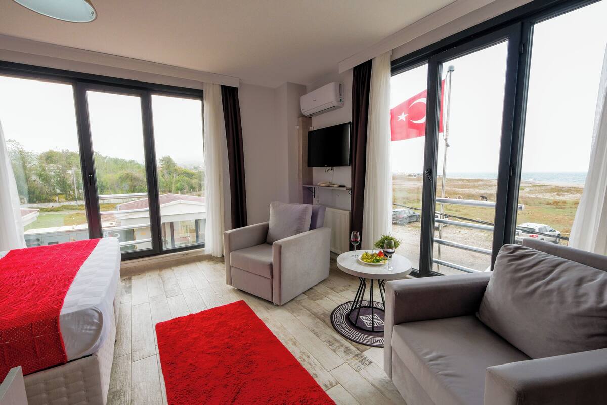 A hotel room with a bed, a grey sofa, a red rug, a small round table with a plant, and a TV mounted on the wall.