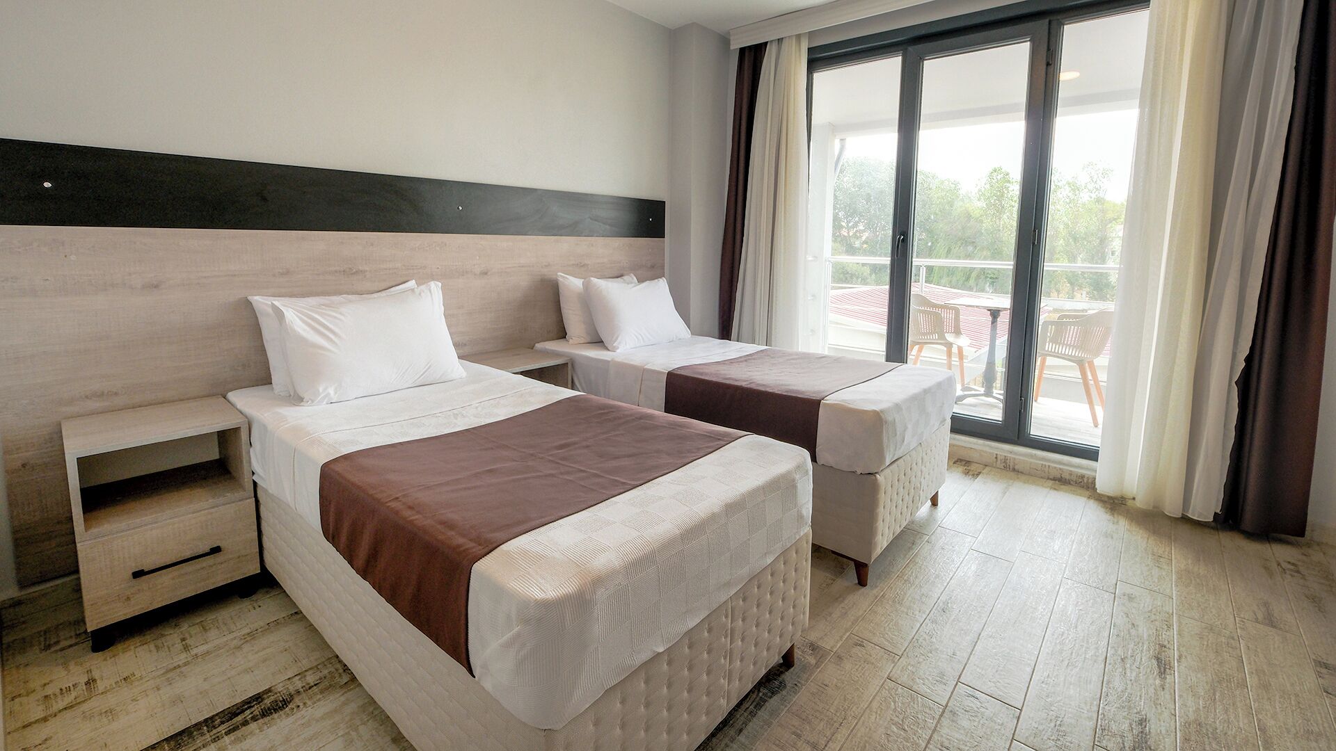 A hotel room with two beds, a wooden headboard, a sliding glass door to the balcony, and a small wooden nightstand.