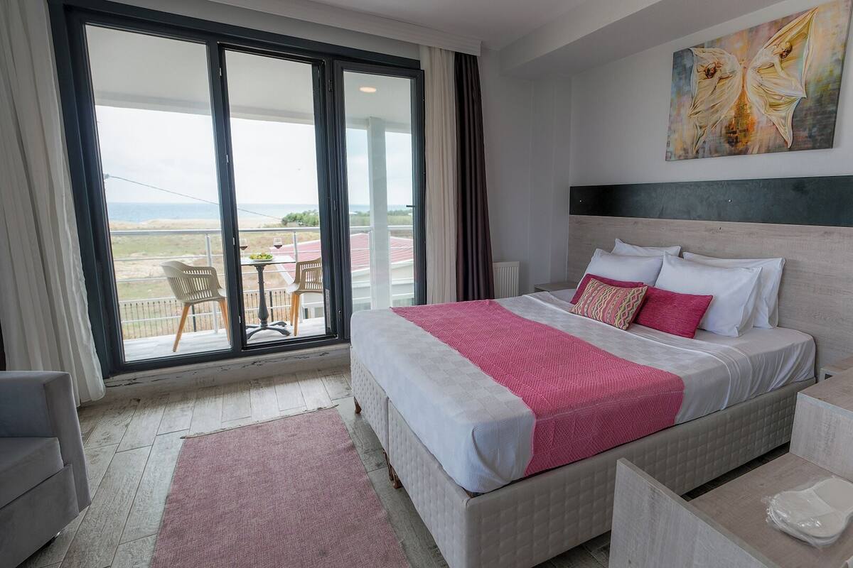 A hotel room with a bed, a desk, a chair, a balcony with a table and chairs, and a painting on the wall.