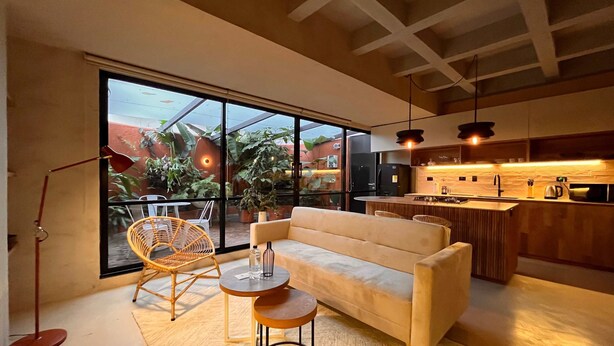 A modern living room with a sofa, two small tables, and a wicker chair. A large window offers a view of a garden. - w* | Charming 1BR Apartment w/ Terrace in Laureles (Medellín)