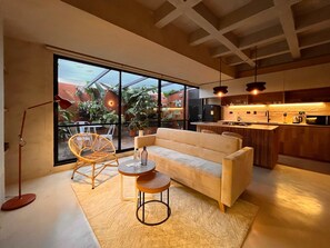Een moderne woonkamer met een bank, twee bijzettafels en een rieten stoel. Een groot raam biedt uitzicht op een tuin.