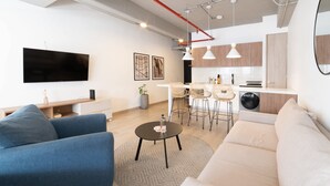 A modern living room with a sofa, blue armchair, and a coffee table. There is a kitchen area with wooden cabinets and a dining table with chairs.