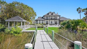 A wooden boardwalk leading to a large house with a balcony, surrounded by lush greenery and palm trees. - Waterfront Home w Patio, Heated Pool, Dock, Beach (St. Augustine)