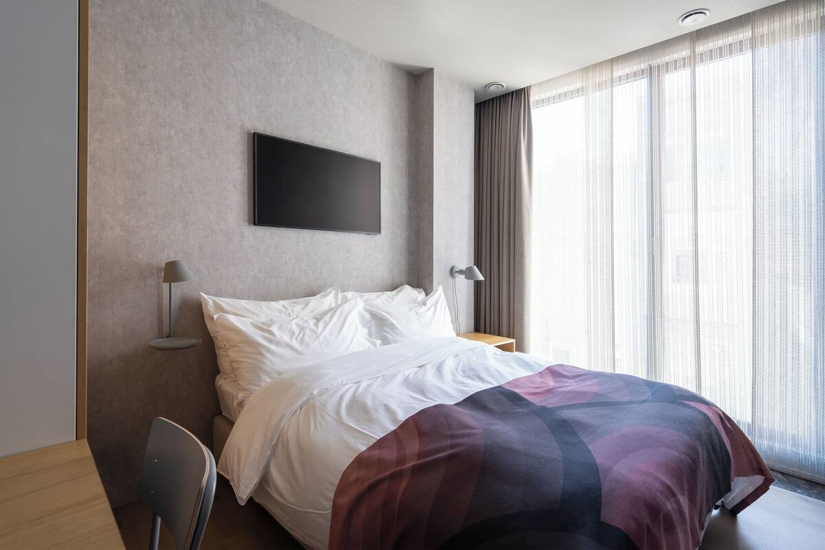 Una habitación de hotel moderna con una cama grande, un televisor de pantalla plana en la pared, un escritorio con una silla y una ventana con cortinas ligeras.
