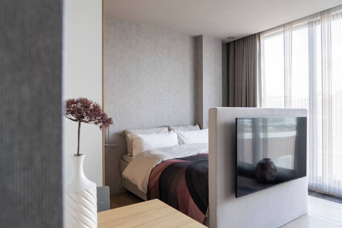 Una habitación de hotel moderna con cama, televisión, jarra y ventana con cortinas.