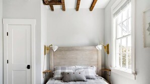 Une chambre à coucher avec un lit, deux lampes de chevet, une fenêtre et des poutres apparentes en bois au plafond.