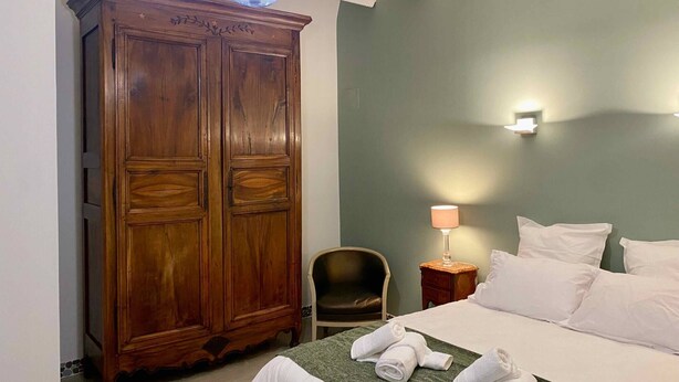 A hotel room with a large wooden wardrobe, a bed with white linens, a green throw blanket, and a nightstand with a lamp.