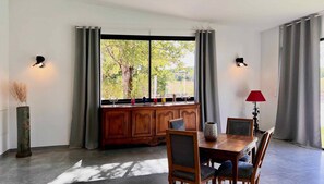 A dining area with a wooden table and chairs, a window with curtains, and a vase with dried plants.