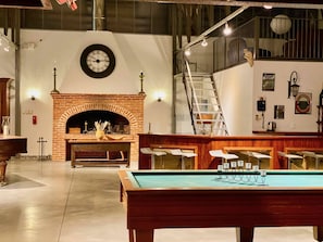 A billiards table in a spacious room with a brick fireplace, a clock, and a staircase leading to an upper level.