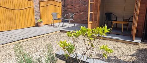 A backyard with a wooden deck, a brick wall, and a wooden door leading to a covered patio area with two chairs and a table.
