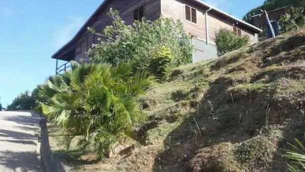 A rustic wooden building with a sloped roof, surrounded by lush greenery and a dirt path. - Greenheart Cottage (Clifton Union island)