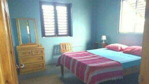 A bedroom with a bed, a dresser, a chair, a window, and a nightstand with a lamp. - Greenheart Cottage (Clifton Union island)