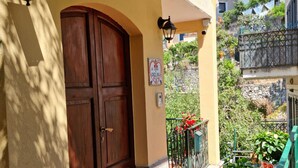 Une porte en bois ornée d’une plaque en forme de cœur, un paillasson, un balcon avec une balustrade et des plantes en pot.