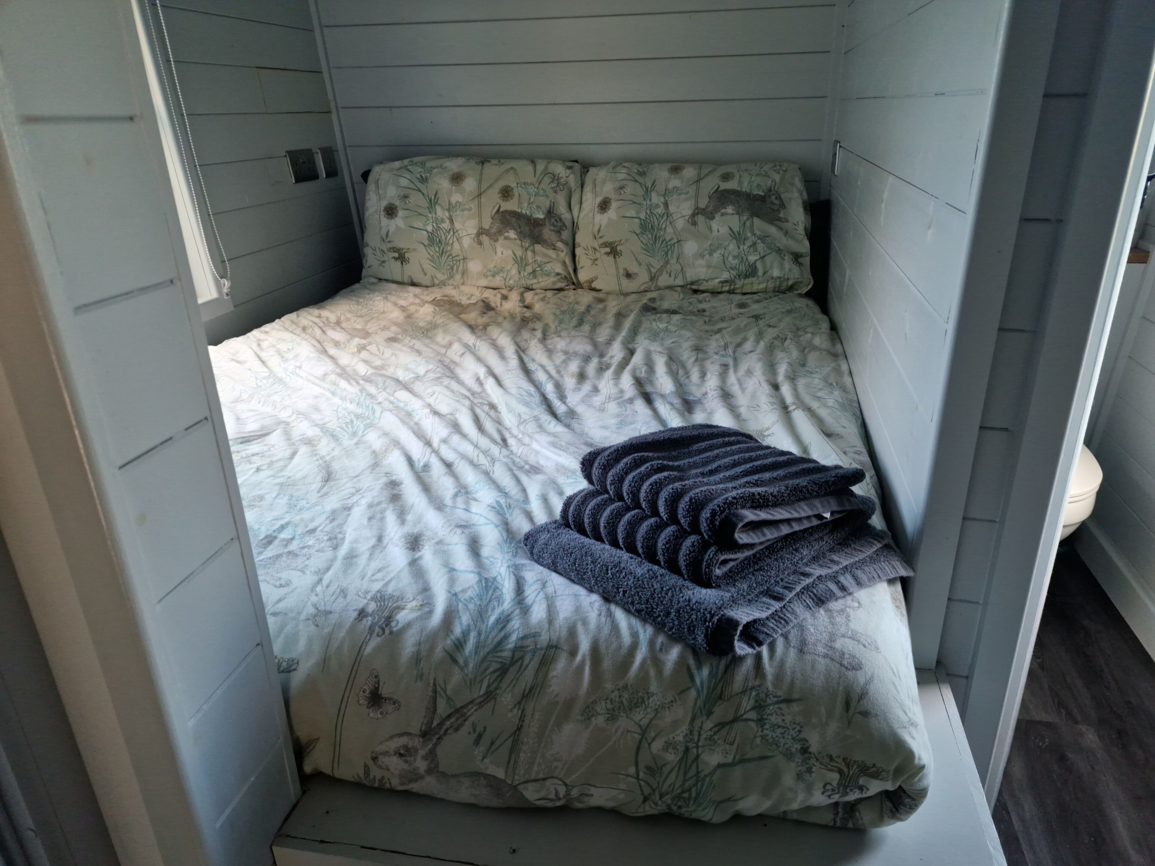 A small, cozy room with a bed featuring a floral and animal print bedspread, a neatly folded dark blue blanket, and a white headboard.