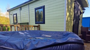 A covered outdoor hot tub with a safety cover, a wooden deck, and a light green building with two windows. - Glamping Hut - Sleeps 2, hot tub & Parking (Welshpool)