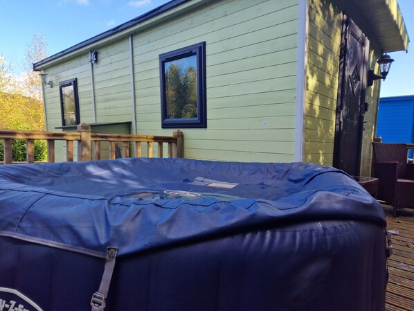 A covered outdoor hot tub with a safety cover, a wooden deck, and a light green building with two windows.