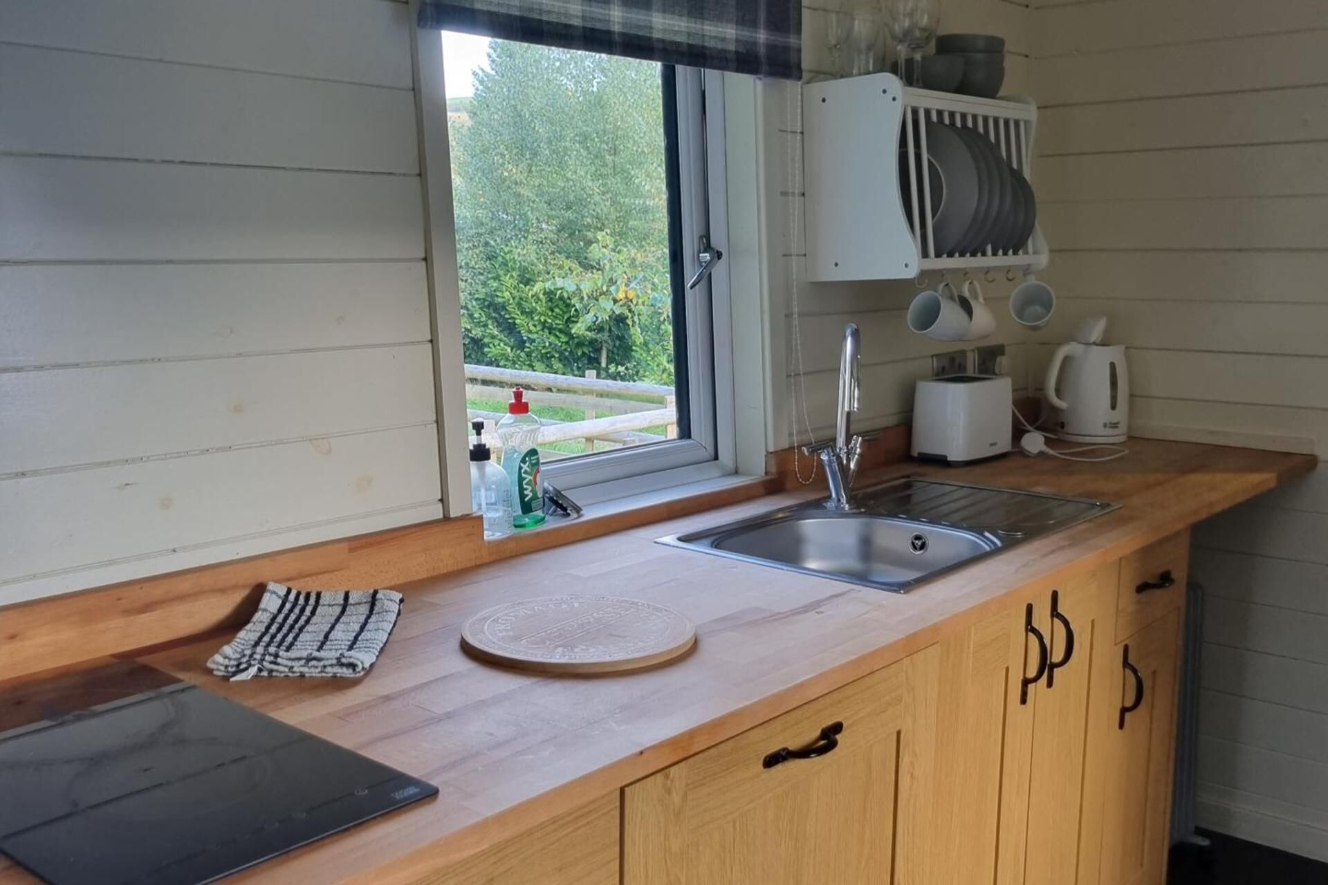 Eine Küche mit einer Holzausziehtafel, einem Spülbecken, einem Fenster mit Blick ins Grüne und einem Geschirrgestell über der Arbeitsfläche.