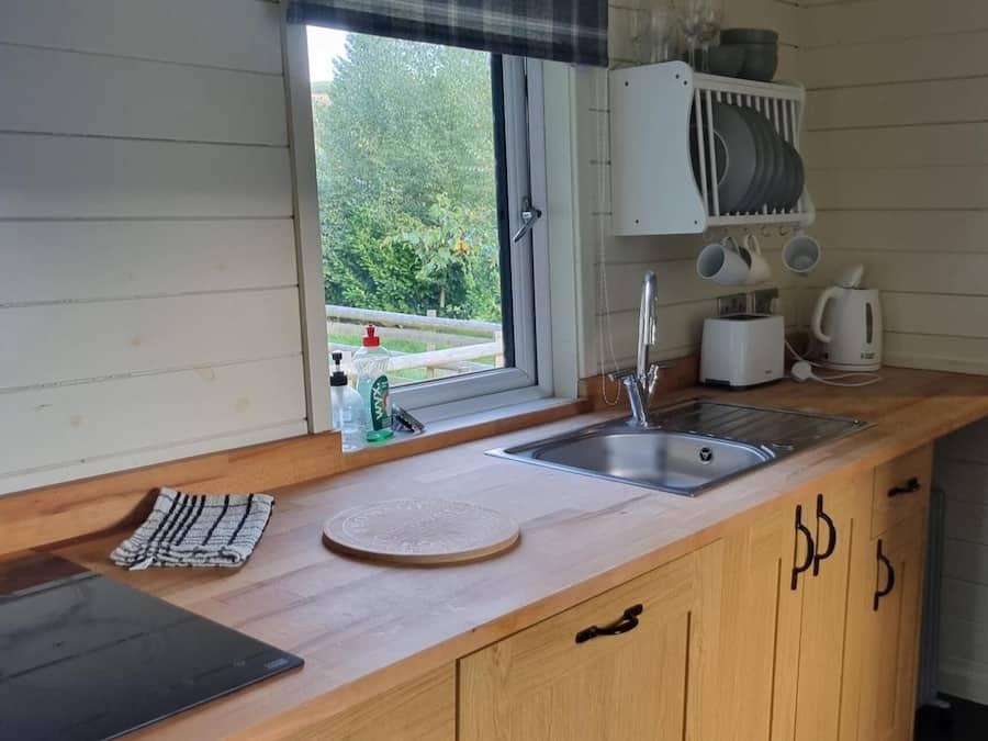Une cuisine avec un plan de travail en bois, un évier, une fenêtre donnant sur un espace verdoyant et un égouttoir à vaisselle placé au-dessus du plan de travail.