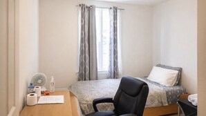 A bedroom with a bed, a chair, a desk with a fan, and a window with curtains. - Henderson Héritage Home (Ottawa)