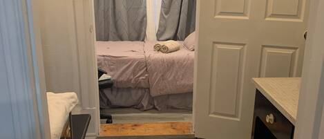 A narrow hallway leading to a bedroom with a bed, a small table, and a door.