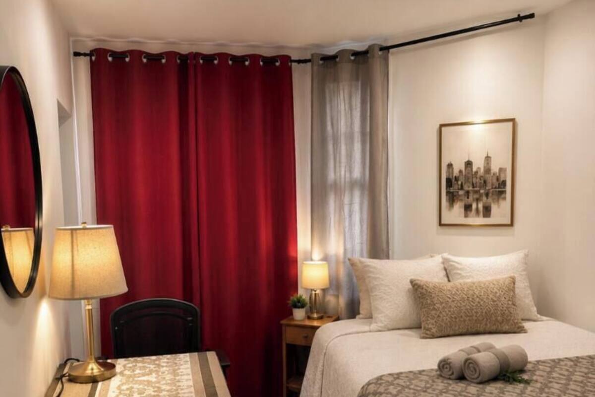 A hotel room with a bed, a desk, a chair, a lamp, a bedside table, and a wall-mounted picture.