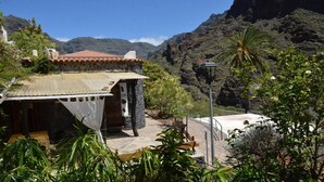 A rustic building with a tiled roof, surrounded by lush greenery and a mountainous backdrop. - Casa Corma (Valle Gran Rey)