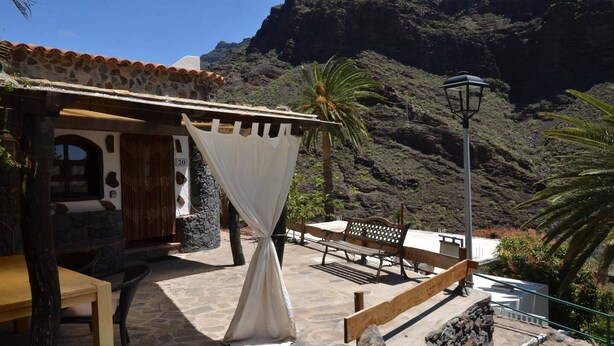 A rustic building with a stone facade, a wooden door, and a number '20' sign. There is a bench and a table outside, with a lamp post nearby. - Casa Corma (Valle Gran Rey)