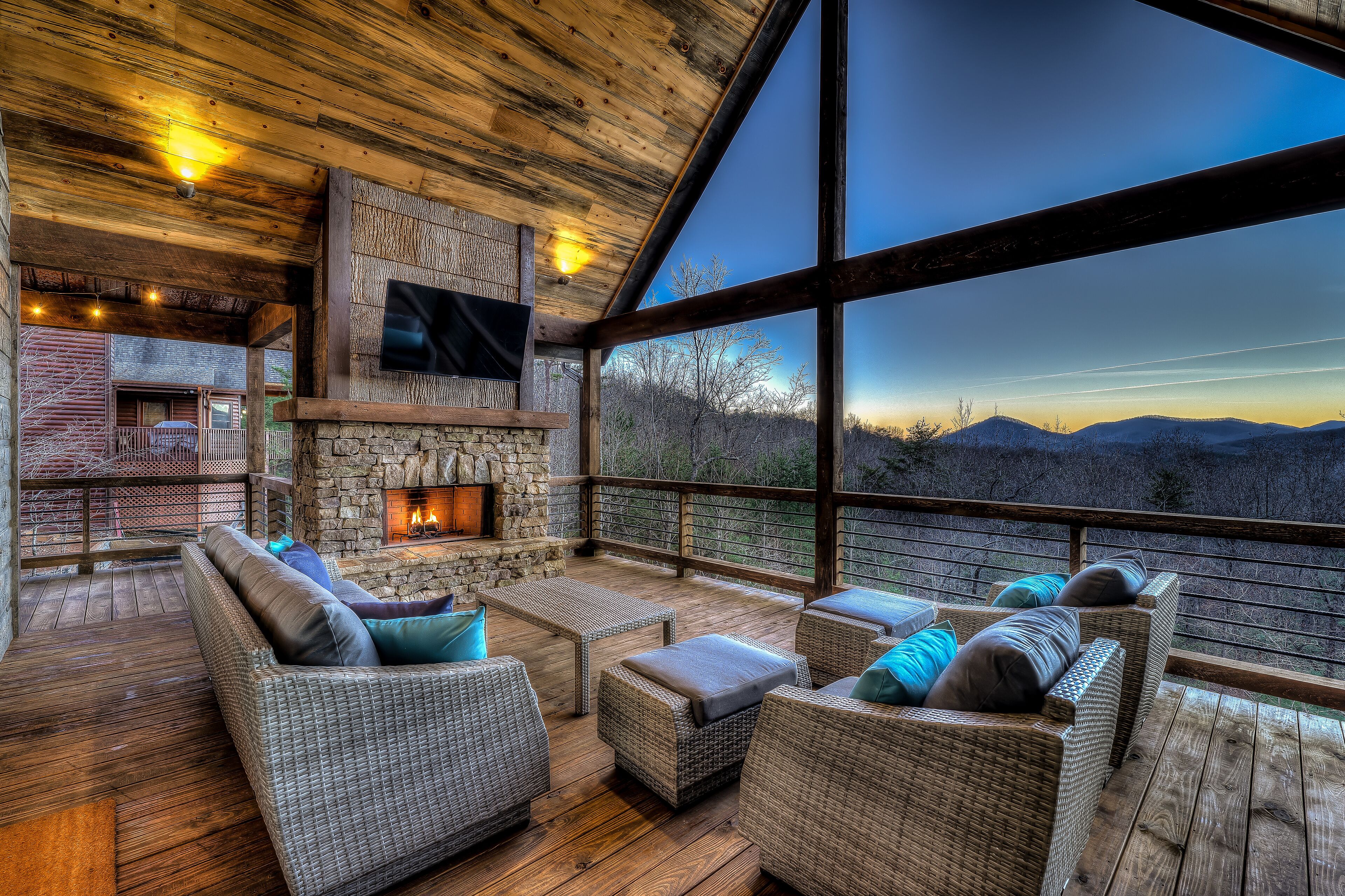 A cozy outdoor seating area with wicker furniture and a stone fireplace, overlooking a mountainous landscape.