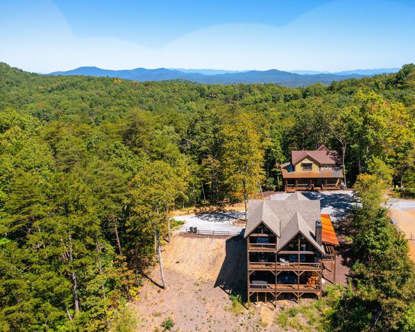 A lodge with a large deck and multiple rooms, surrounded by dense forest and mountains in the background.