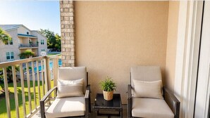 Ein Balkon mit zwei Korbstühlen, einem kleinen Tisch, einem Blumentopf und Blick auf einen Swimmingpool und Gebäude.