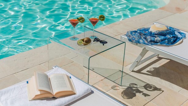 Une table en verre avec un livre, une paire de lunettes et deux boissons, près d’une piscine.