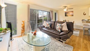 A modern living room with a glass coffee table, a leather sofa with decorative pillows, a dining area with a bar stool, and a large sliding glass door leading to a balcony.