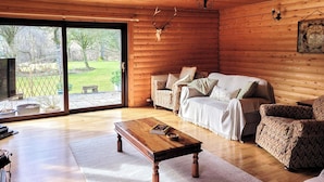 A cozy cabin interior with a wooden ceiling, a sofa, a patterned armchair, a wooden coffee table, and a large sliding glass door leading to a garden. - 3 bedroom accommodation in Enochdhu, near Pitlochy (Enochdhu, near Pitlochy)