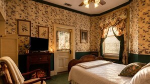 A bedroom with floral wallpaper, a bed, a dresser, a television, and a window with curtains. - Bailey Inglish Suite • Private Balcony (Bonham)