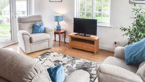 A living room with a sofa, a television on a wooden stand, a lamp, and a window with a view of greenery. - The Coach House - UK41070 (Wootton, near Ryde)