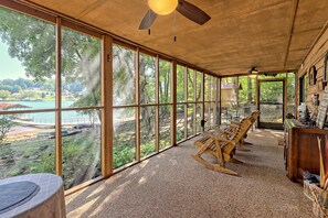 Habitación con ventanas amplias que ofrecen vista a un lago y un bosque. Se observan sillones de madera con movimiento y una mesa con mantel blanco.