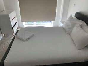 En pænt redt seng med hvide sengetøj, en grå sengegavl og et vindue med skodder.