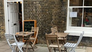 An outdoor seating area with wooden tables and chairs, a chalkboard sign advertising fresh bread and pastries, and a brick wall backdrop. - Luxury 2-Bed High-rise Apartment with beautiful view near Excel, &O2. Next to CR (London)