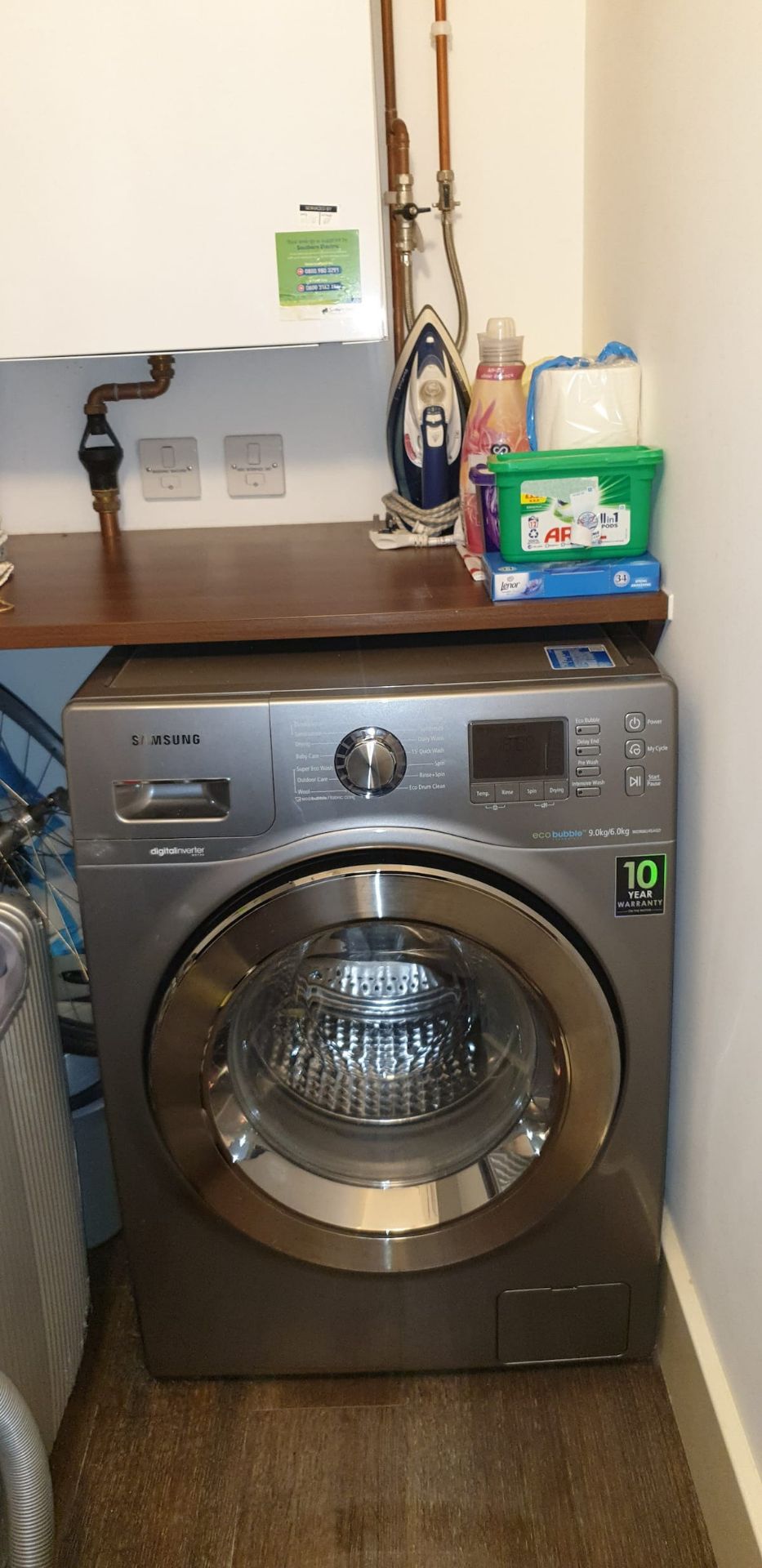 A Samsung digital inverter washing machine with a 10-year warranty, placed in a laundry room with a wooden counter, a water heater, and various laundry supplies.