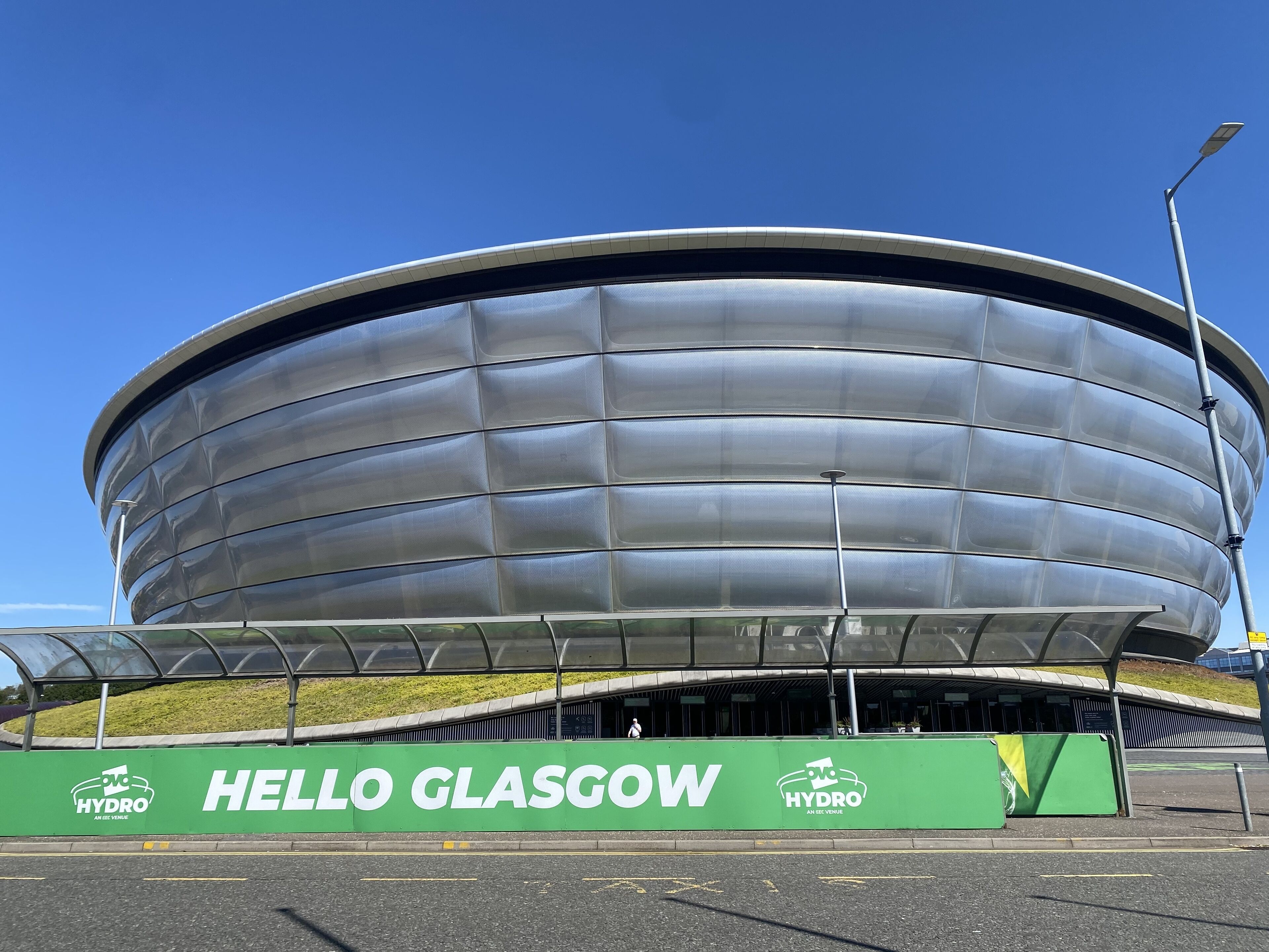 A modern stadium with a curved design and a green 'HELLO GLASGOW' sign in front.