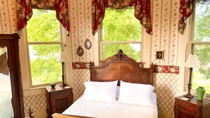 Ein Schlafzimmer mit einem hölzernen Bett, zwei Nachttischen, einem Fenster mit floralem Vorhang und Blick ins Grüne.